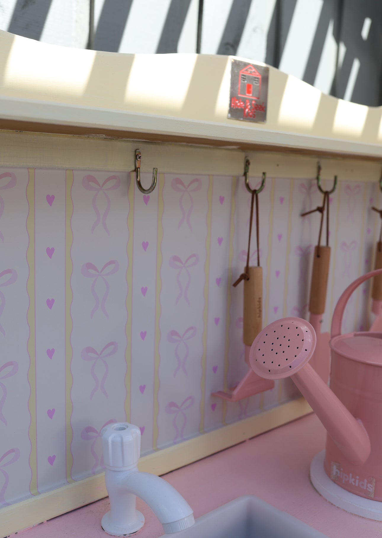 Pink watering can and white faucet on a pink countertop with a decorative wall in the background.